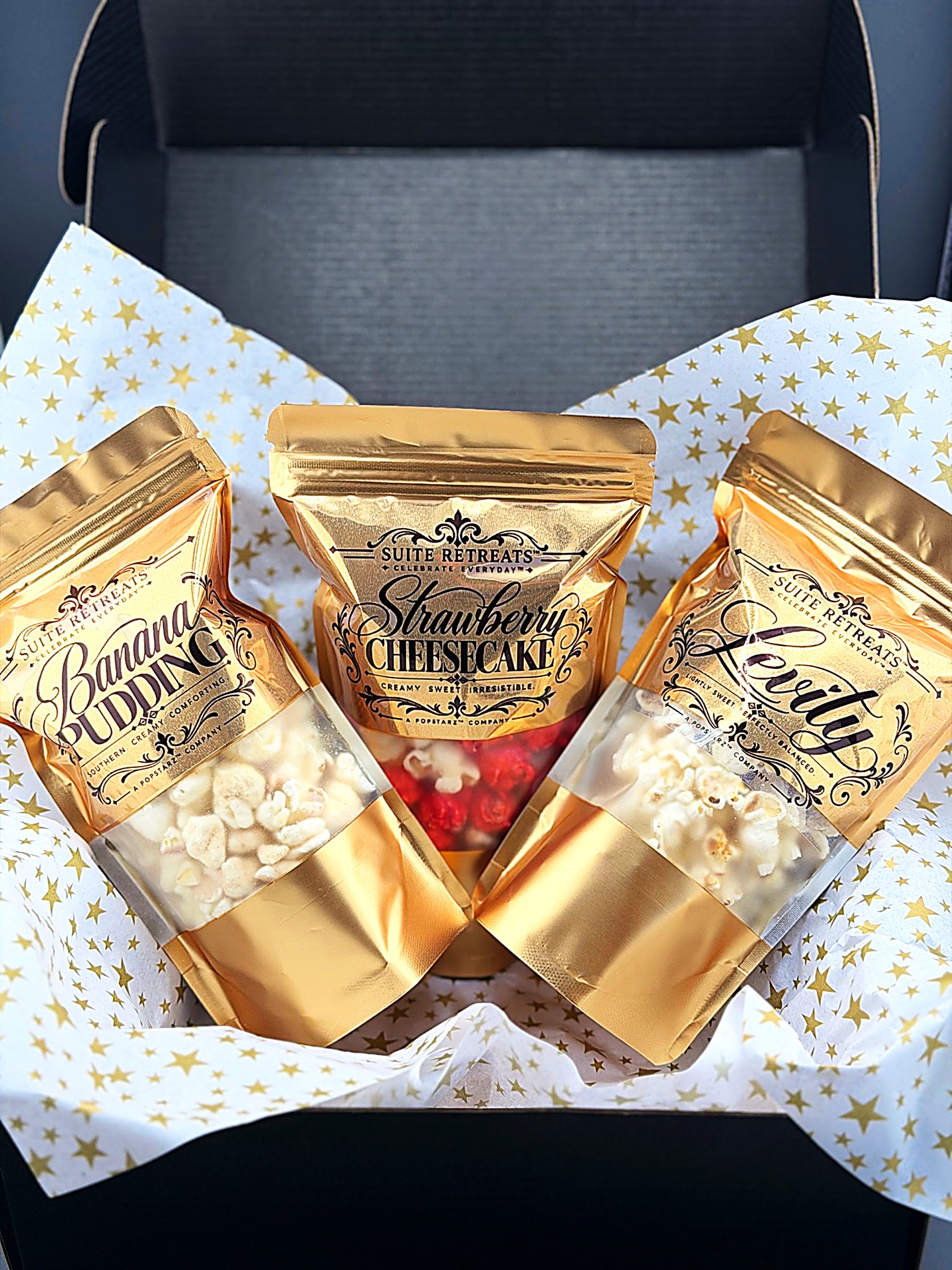The signature Suite Retreats black gift box, open, with gold pouches of Banana Pudding, Strawberry Cheesecake and Levity popcorn nestled in gold-star tissue