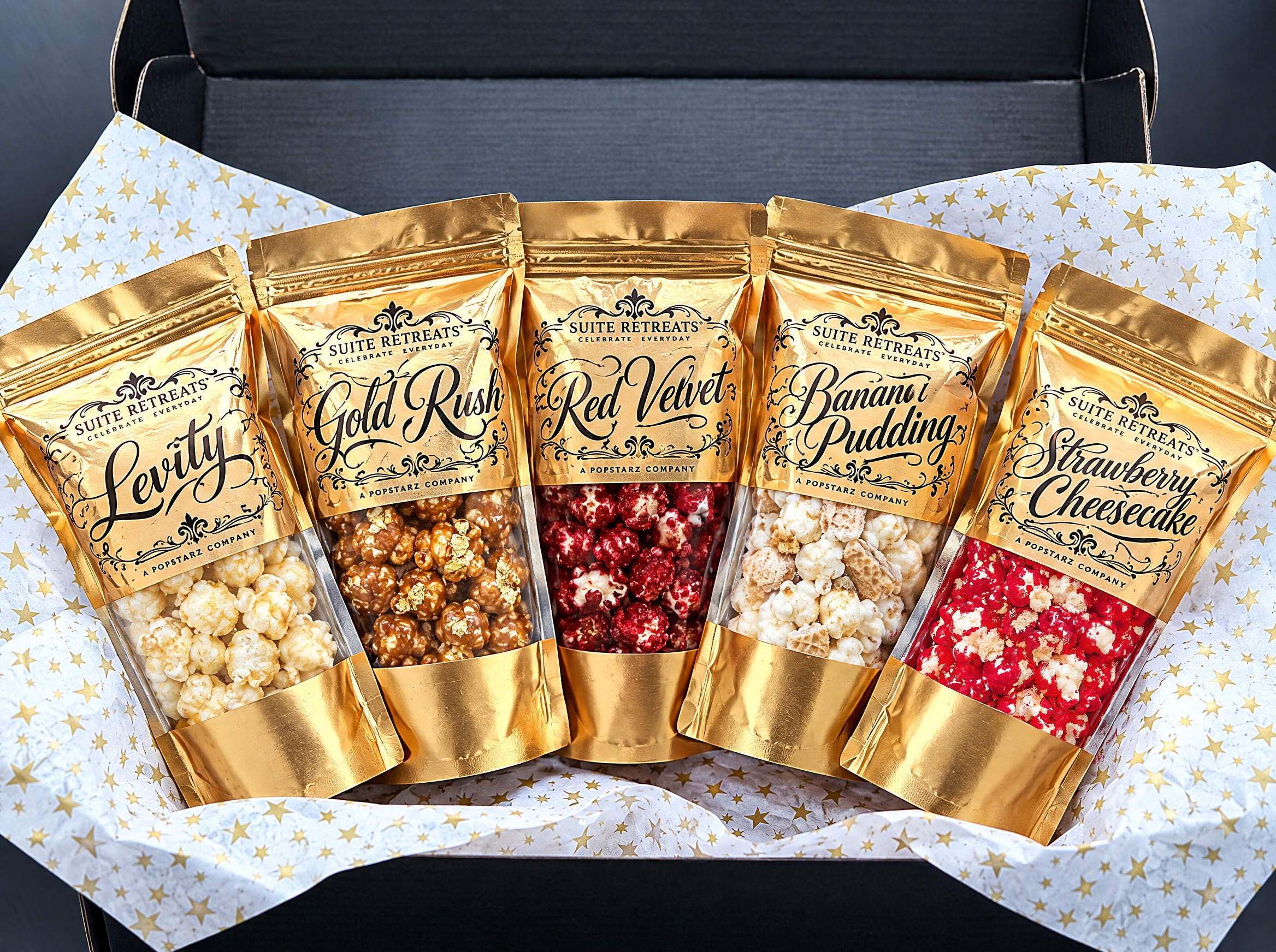 All five Suite Retreats gold pouches — Levity, Gold Rush, Red Velvet, Banana Pudding and Strawberry Cheesecake — fanned inside the open black gift box on gold-star tissue